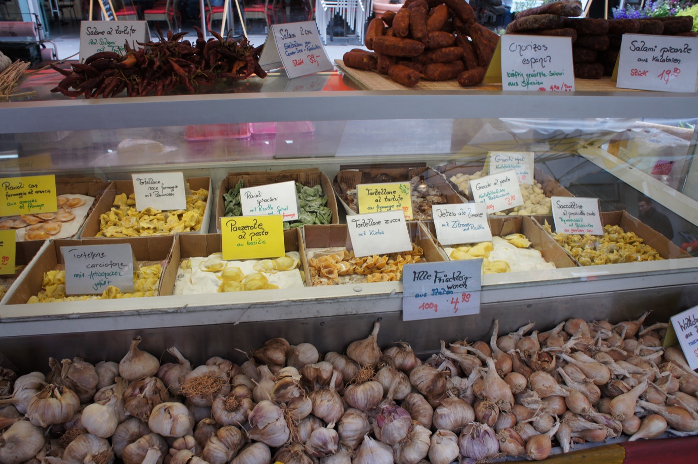 イタリア食材の屋台。上段には吊るされたサラミやスペインのチョリソー、赤唐辛子。中段のガラスケースには、かぼちゃ(Kürbis)やトリュフ(t
